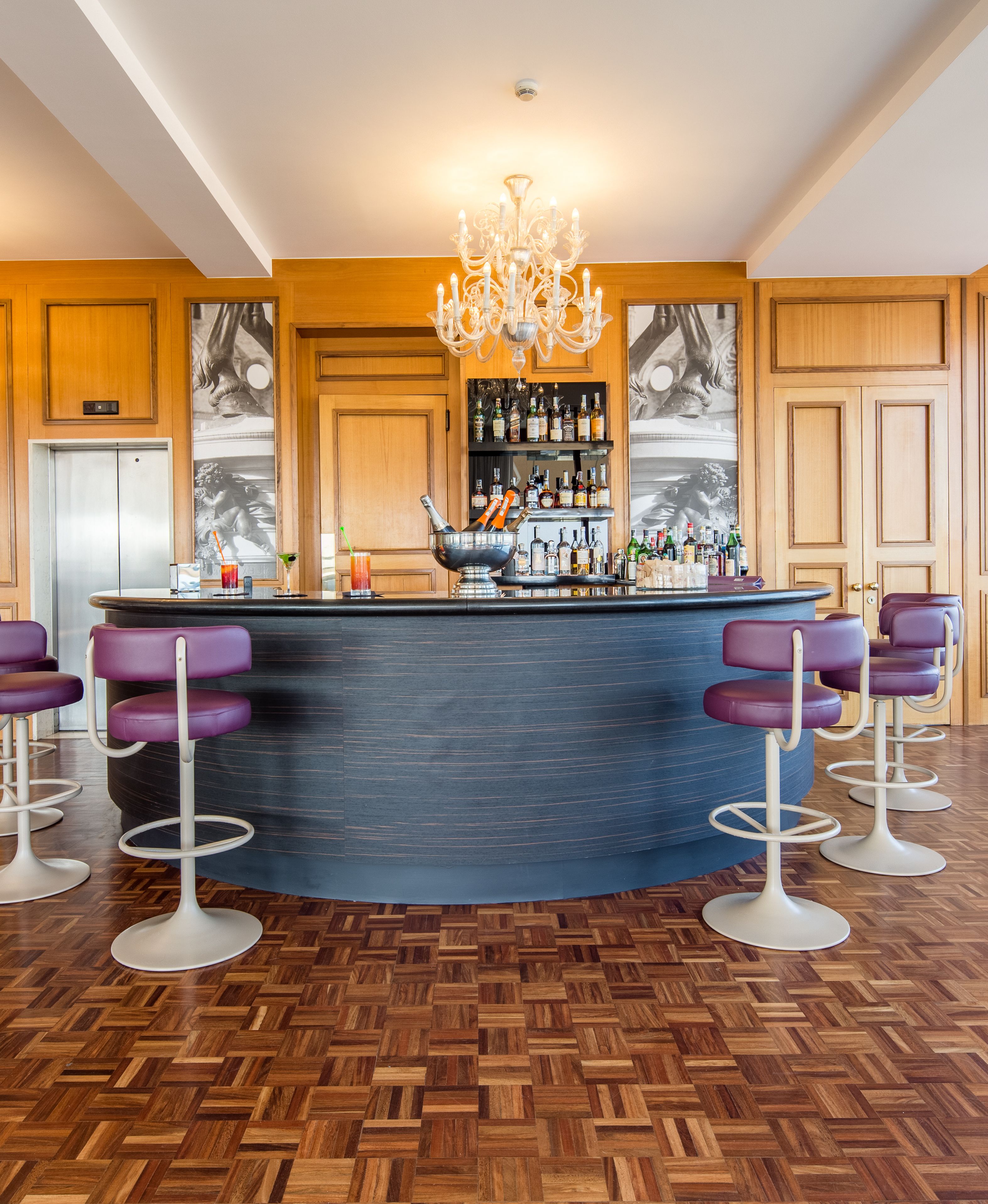 A bar area with a curved counter, purple bar stools, a chandelier, and a wooden floor.