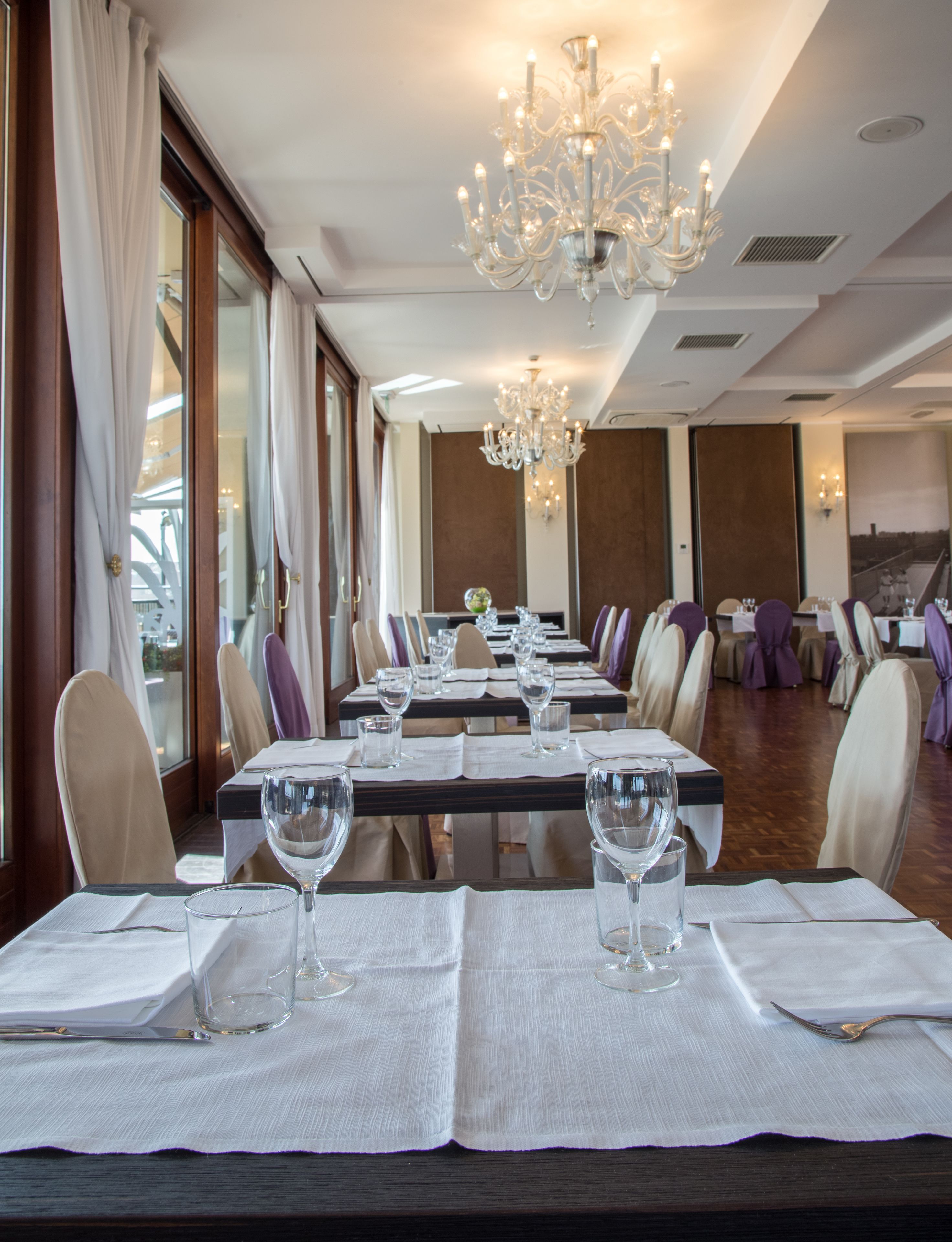 A dining area with tables set for a meal, a chandelier, and large windows.