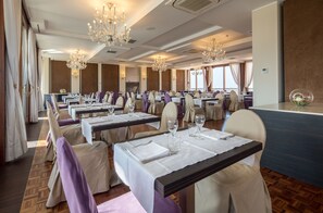 A formal dining room with multiple tables set for service, elegant chandeliers, and a view of the outside through large windows.