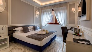 A modern hotel room with a marble floor, a bed with a dark blue headboard, a bedside table with a lamp, and a desk with a mirror and a vase of flowers.