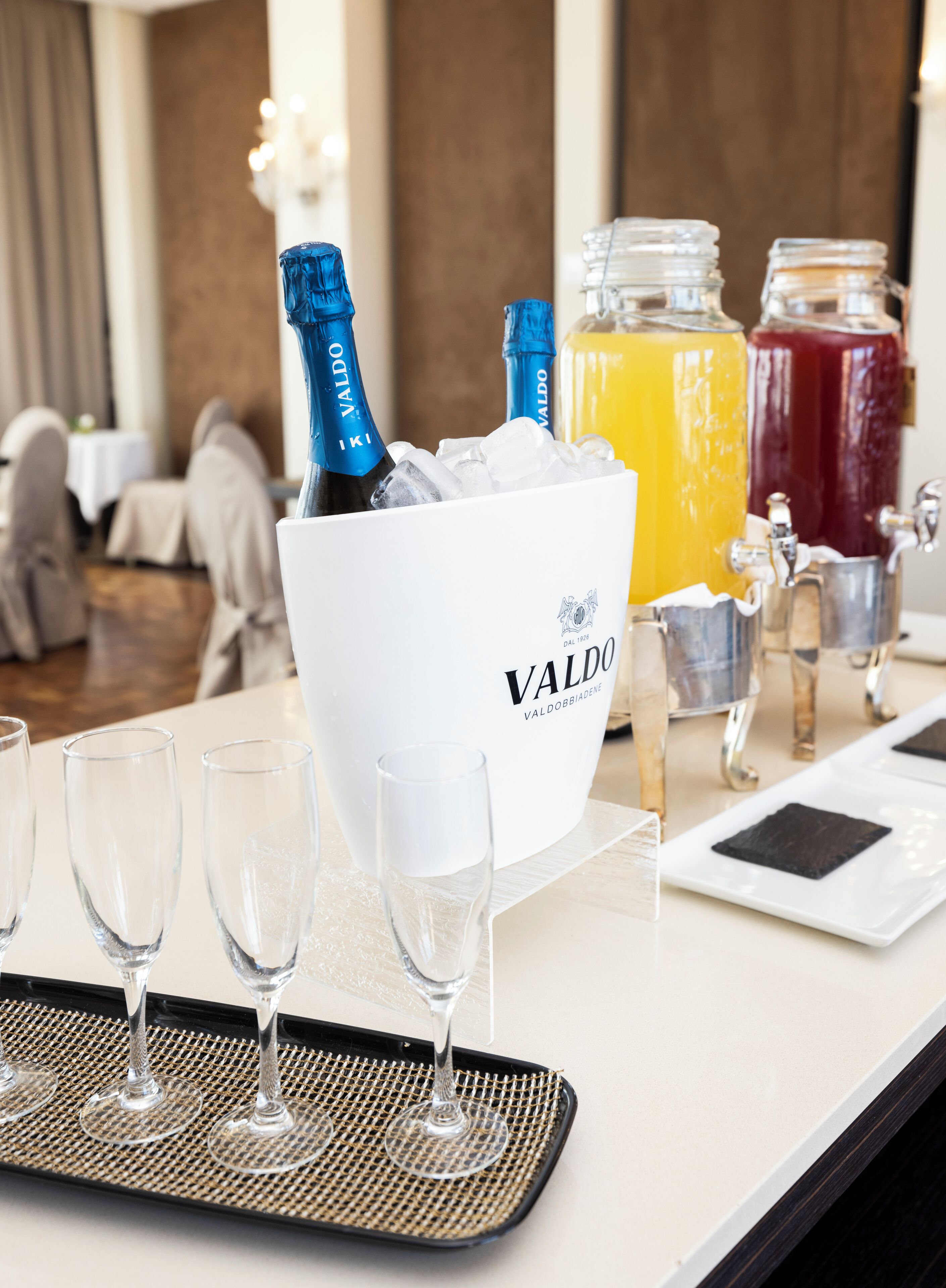A bar area with a white bucket containing champagne bottles, a large pitcher of orange juice, and a red drink. There are empty glasses on a tray in front of the bucket.