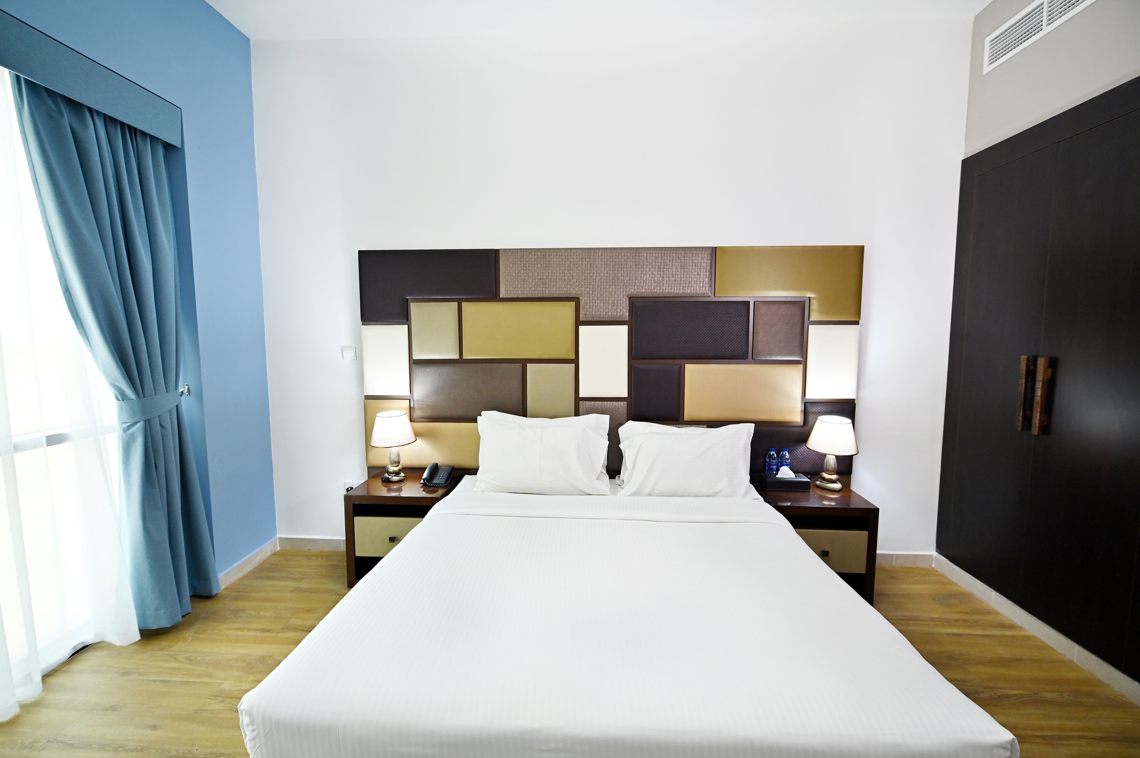 A hotel room with a large bed, two bedside tables with lamps, a headboard with a geometric pattern, a wardrobe, and a window with curtains.
