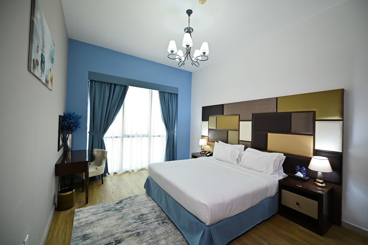 A hotel room with a large bed, a nightstand with a lamp, a desk with a chair, and a window with blue curtains.
