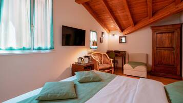 Ein Schlafzimmer mit Holzbalkendecke, einem Bett mit grüner Bettwäsche, einem Schreibtisch mit Stuhl, einem Kleiderschrank und einem Fernseher an der Wand.