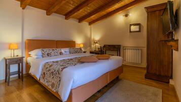 Une chambre avec un plafond en bois, un lit recouvert d’une literie à motifs floraux, des lampes de chevet, une armoire en bois et un radiateur.