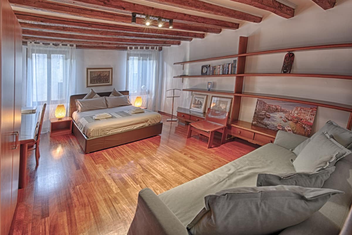 Een moderne slaapkamer met een houten vloer, een bed met hoofdeinde, een bank, een bureau en planken met boeken en foto's.