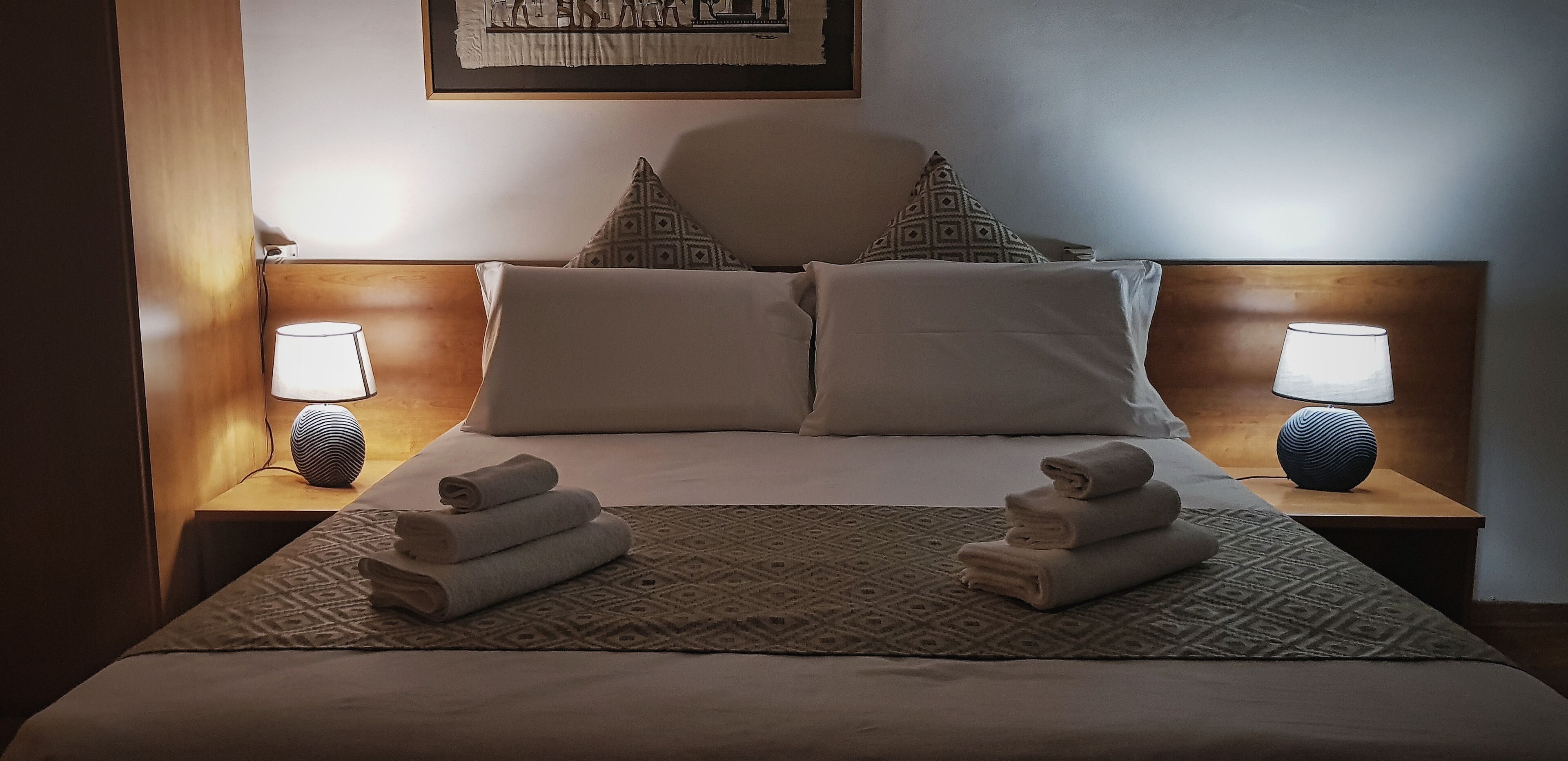 Een net opgemaakt bed met twee kussens, twee nachtlampjes en twee opgerolde handdoeken op een kamerbreed tapijt.