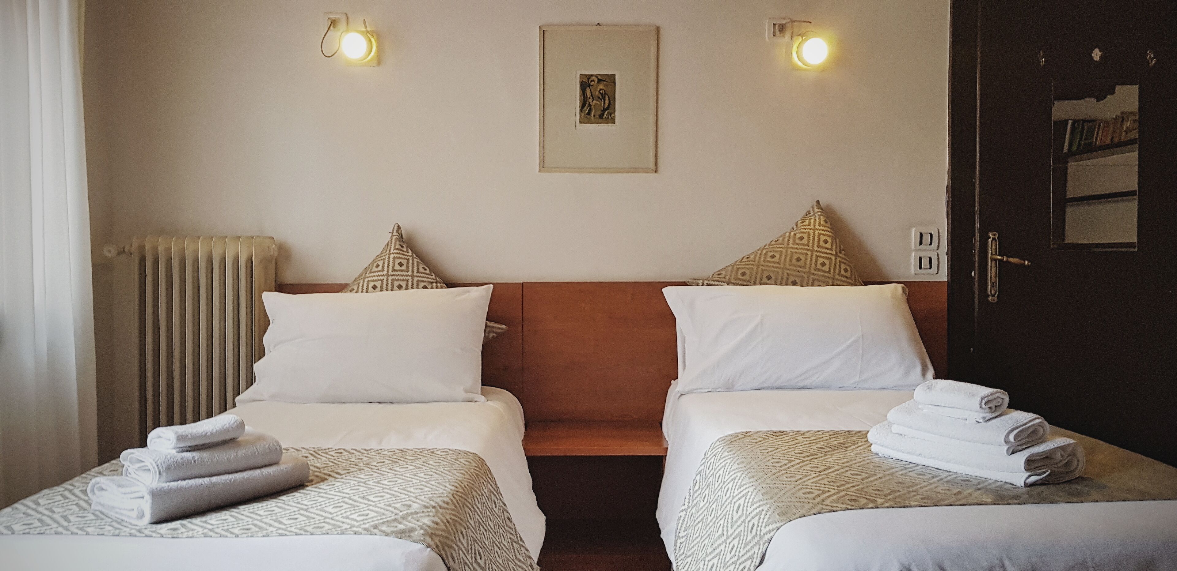 A hotel room with two beds, each with a patterned bedspread, a framed picture on the wall, and two wall-mounted lights.