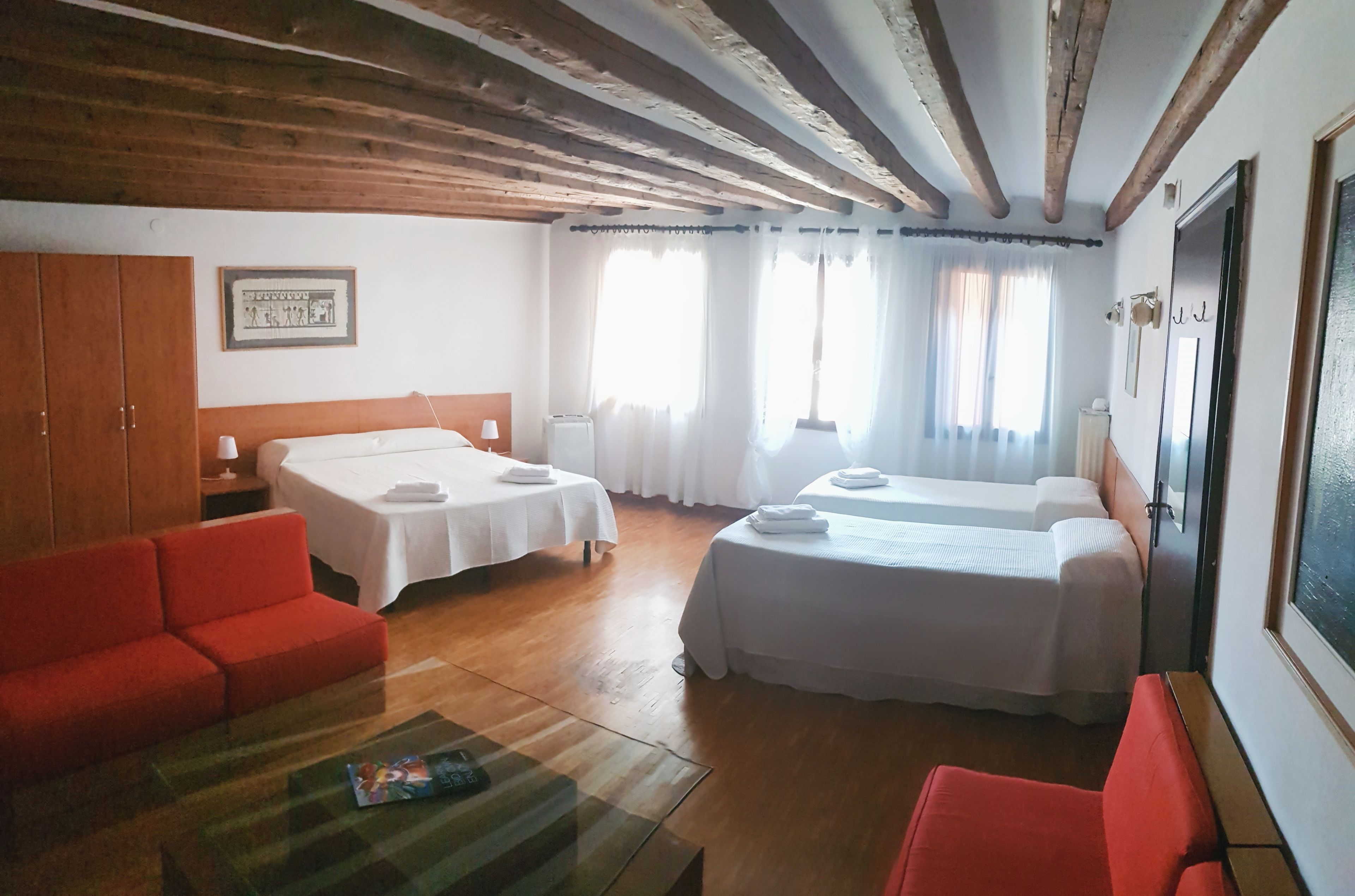 Habitación de hotel con dos camas, un sofá rojo, una mesa de centro de vidrio, vigas de madera en el techo y un cuadro enmarcado en la pared.