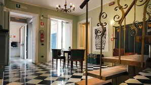 Een hotel lobby met een zwart-wit geruit vloerkleed, een trap met versierde metalen leuningen en een kroonluchter.