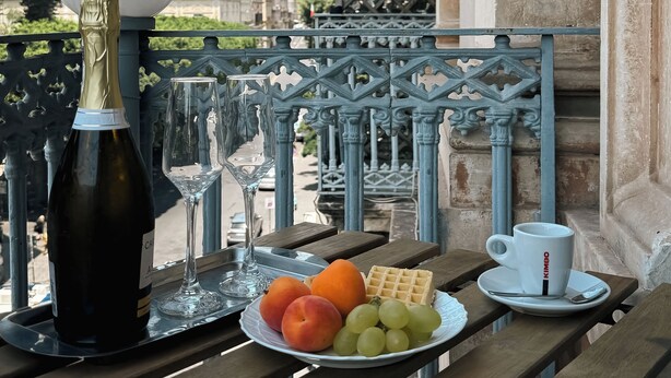 Een tafel met een fles wijn, een glas, een bord met fruit en een koffiekop op een balkon met uitzicht over de stad.