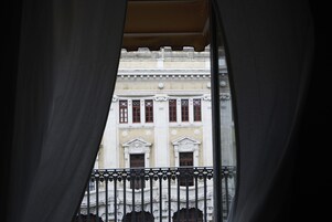 Vista attraverso una finestra con ringhiera di balcone, su un edificio con architettura classica e numerose finestre.