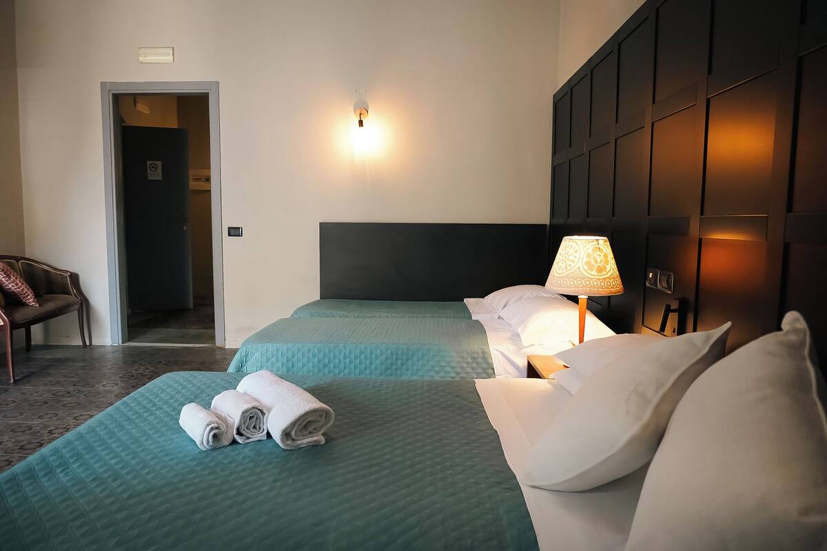 A hotel room with two beds, a nightstand with a lamp, a chair, and a door to another room.