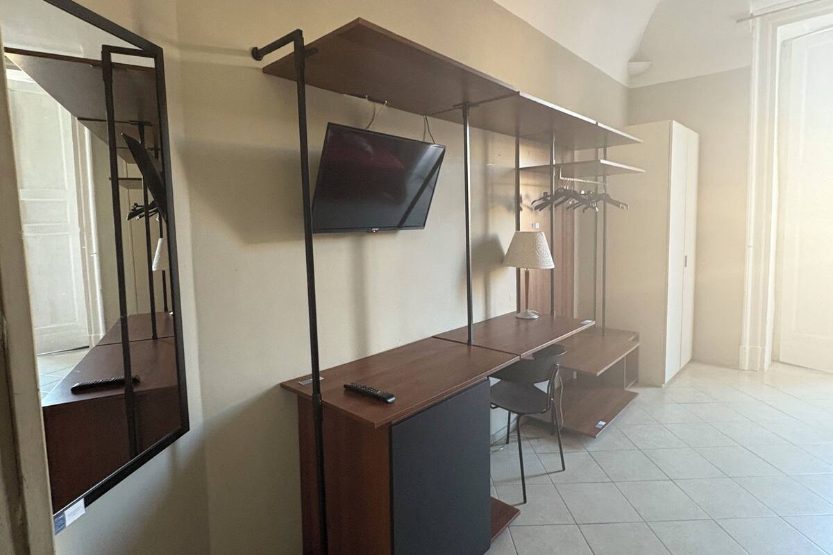 A spacious room with a long wooden desk, a mounted flat-screen TV, and a mirror on the left wall.