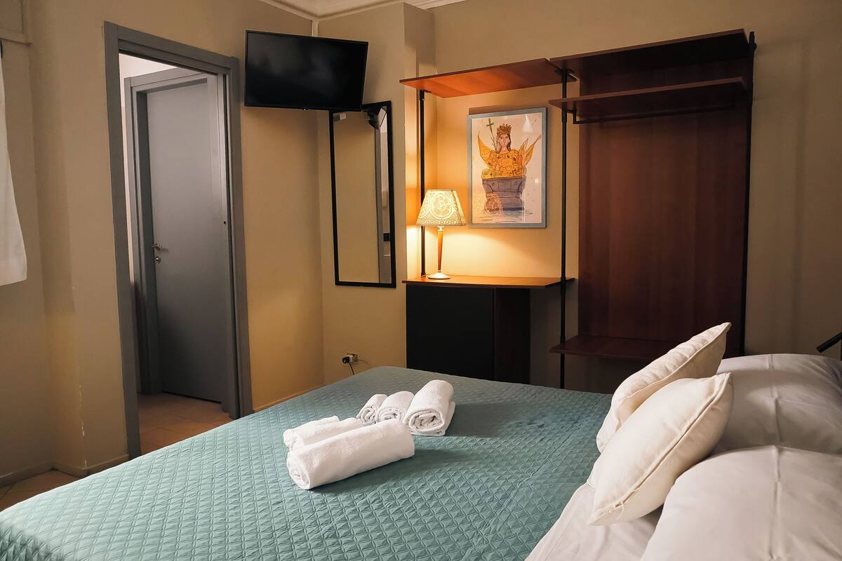 A hotel room with a bed, a TV mounted on the wall, a desk with a lamp, and a painting on the wall.