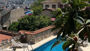 Piscina con sillones de descanso, pared de piedra y vista a un paisaje urbano y montañas.