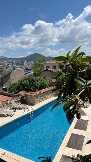 A swimming pool with lounge chairs, a stone wall, and a view of a cityscape and mountains.