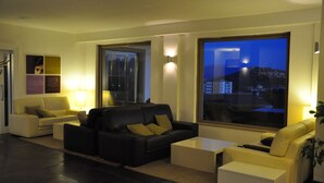 A modern living room with a black leather sofa, a yellow sofa, and a white coffee table. There are two wall-mounted lamps and a large window with a city view. - Hotel Sandalia (Nuoro)
