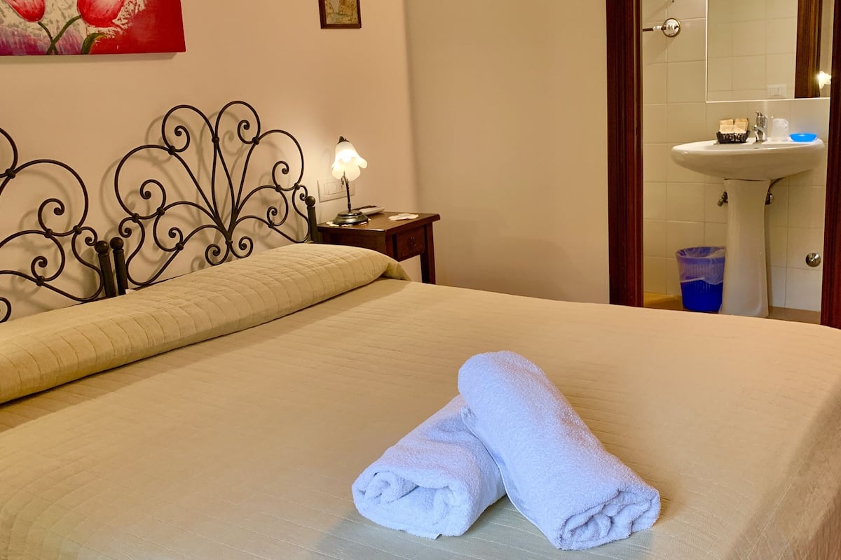 A neatly made bed with two rolled towels, a bedside table with a lamp, a painting on the wall, and a bathroom visible through an open door.