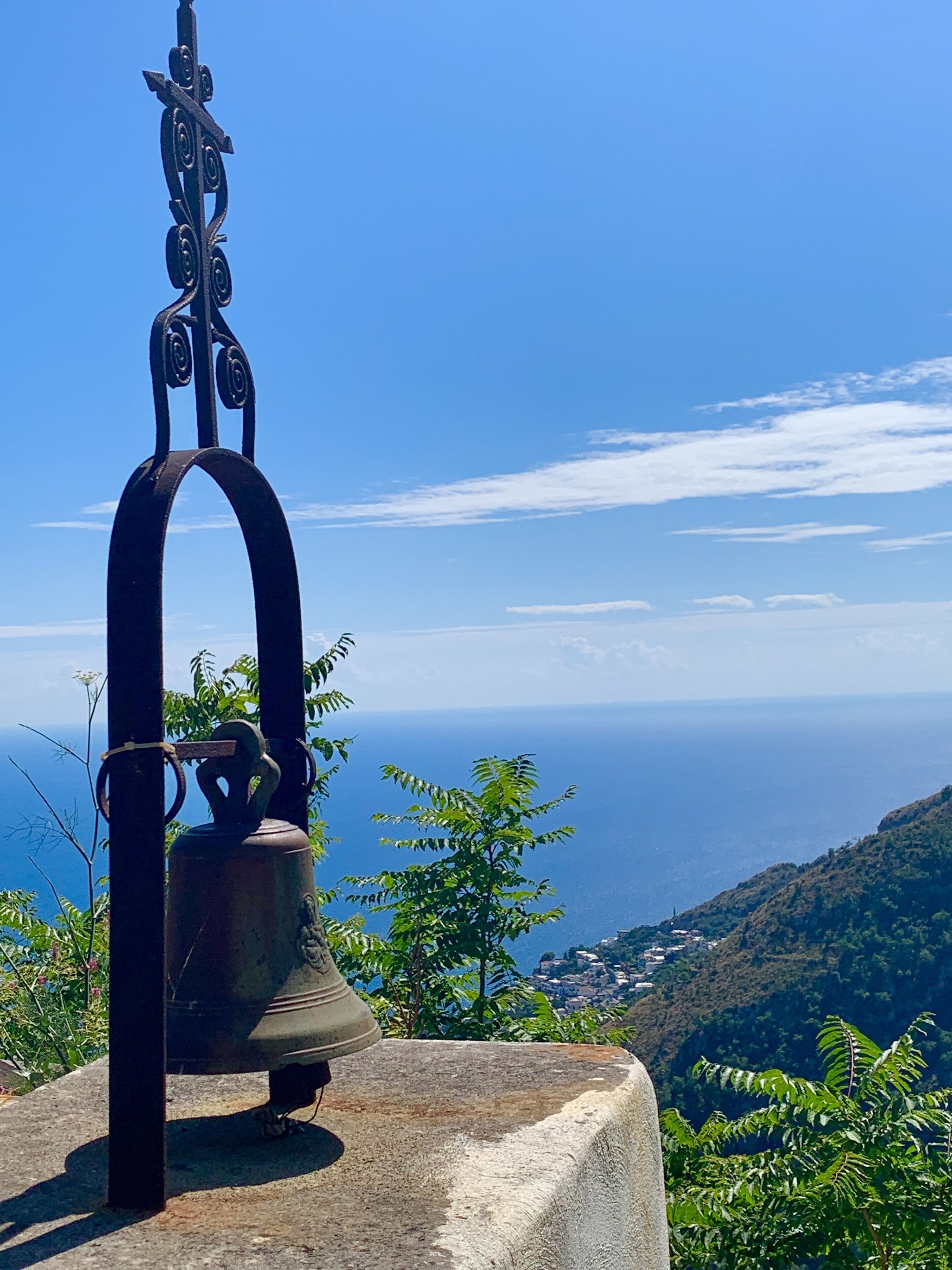Una campana sobre un pedestal de piedra con estructura metálica, con vistas a un paisaje costero con colinas y cielo despejado.