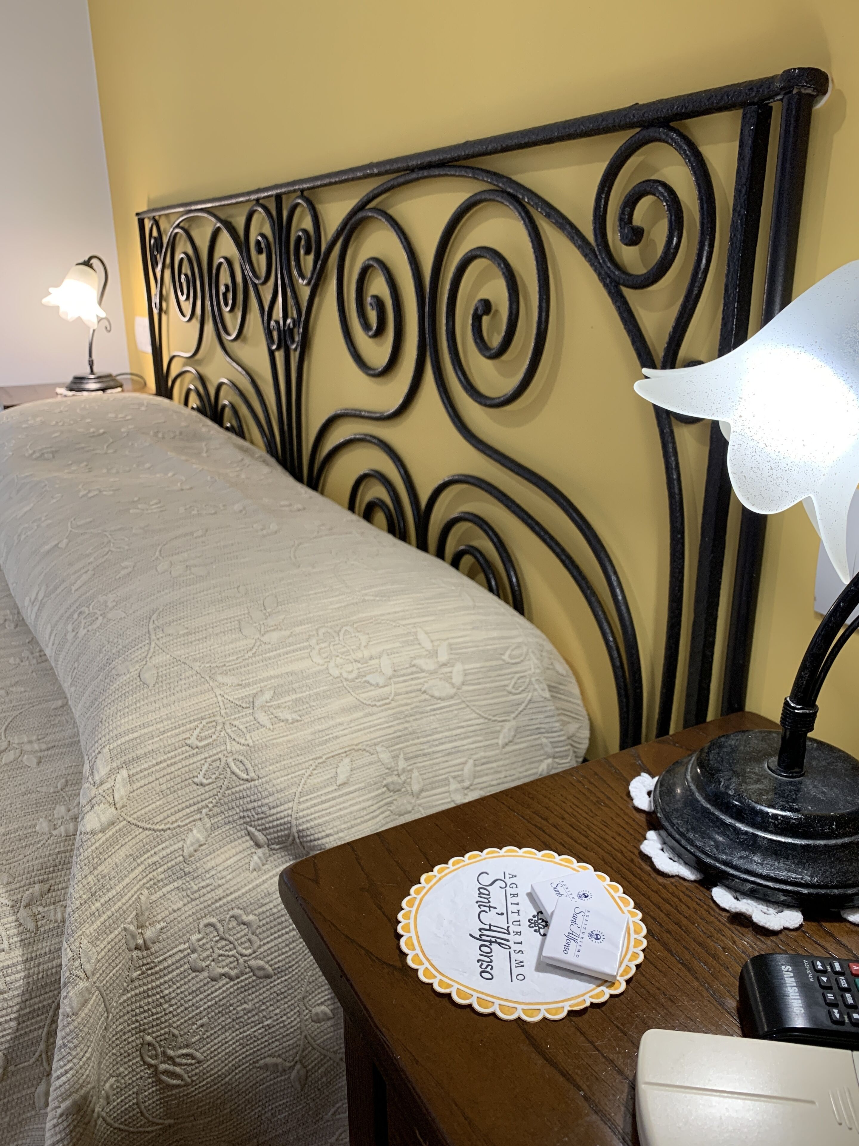 A neatly made bed with a decorative headboard, a bedside table with a lamp, a remote control, and a placemat with a name.