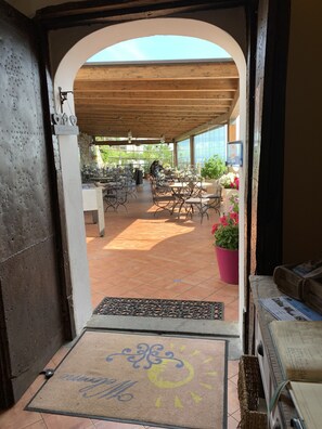 Una puerta abierta que da a un patio techado al aire libre, con mesas y sillas de madera, piso de baldosas y un felpudo.