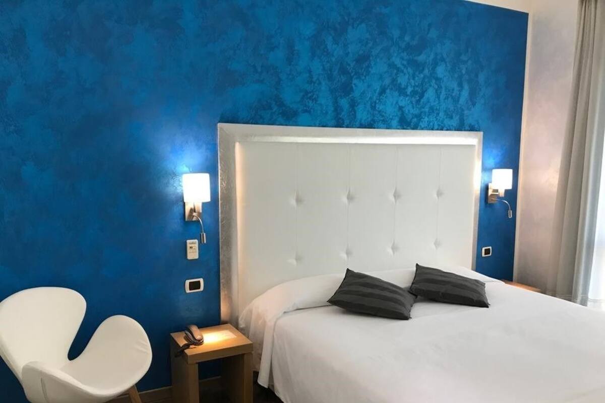 A hotel room with a white bed, a white chair, and a small bedside table.