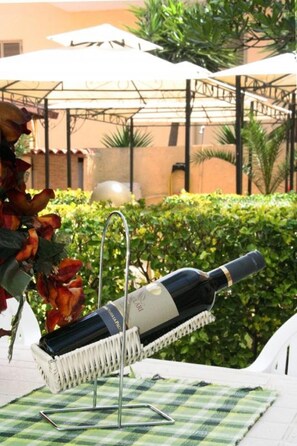 Una botella de vino sobre un soporte de metal con un mantel de cuadros debajo, ubicada al aire libre con una sombrilla de patio y follaje en el fondo.