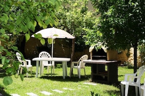 Un patio exterior con sillas y mesas de plástico blanco, una parrilla pequeña y un paraguas decorativo.