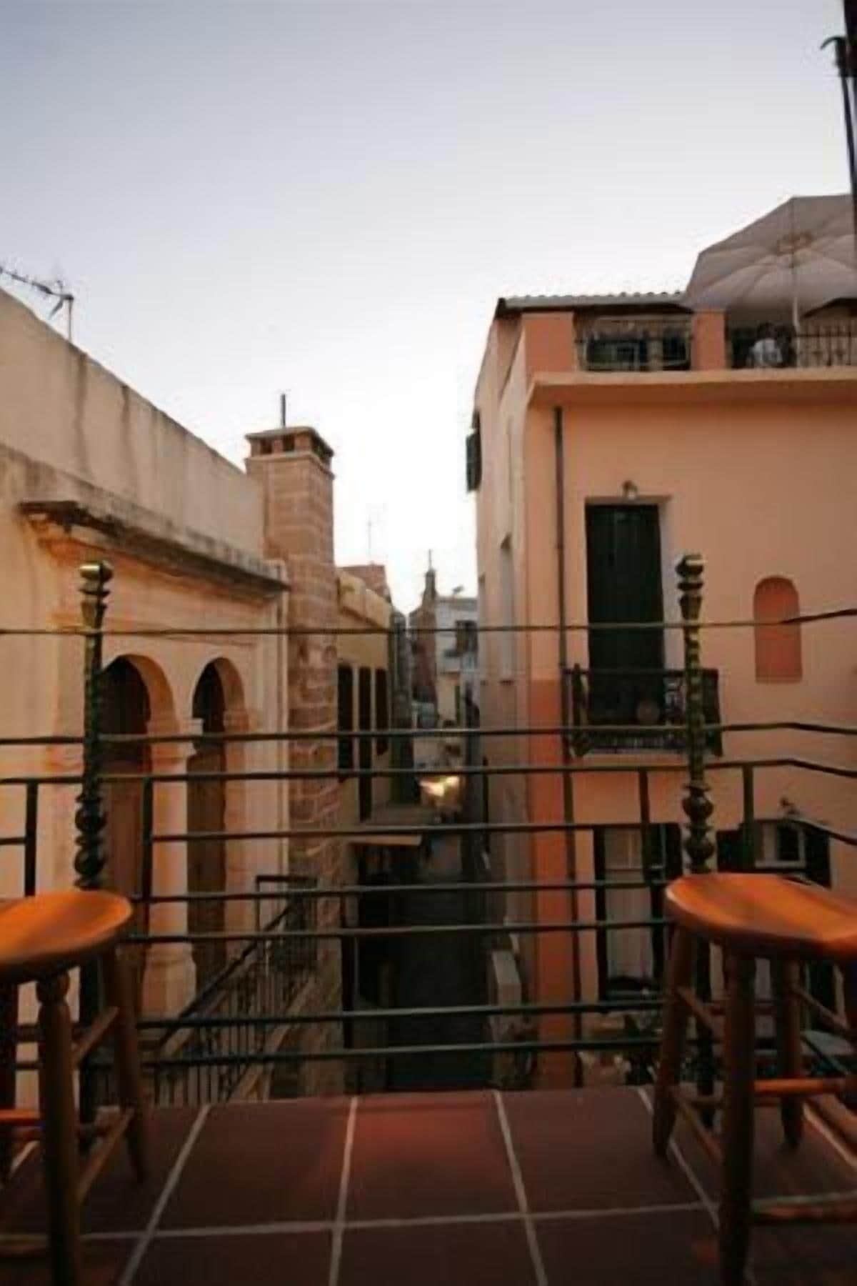 Un estrecho callejón flanqueado por edificios, una baranda metálica y dos taburetes de madera.