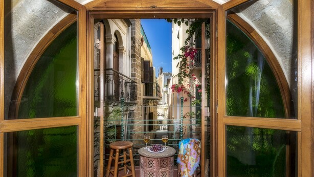 A narrow alleyway with a wooden table and two stools, a hanging plant, and a view of old buildings through arched windows. - Ifigenia Rooms & Studios (Chania)