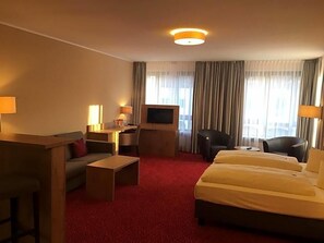 A hotel room with a red carpet, a sofa, a bed, a desk with a TV, and a chair.