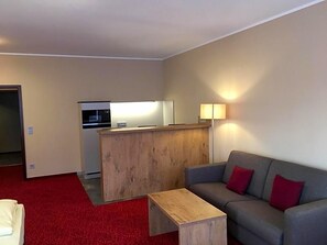 Una habitación con un sofá gris, una mesa de centro de madera, alfombra roja, una kitchenette y un mostrador de recepción de madera.