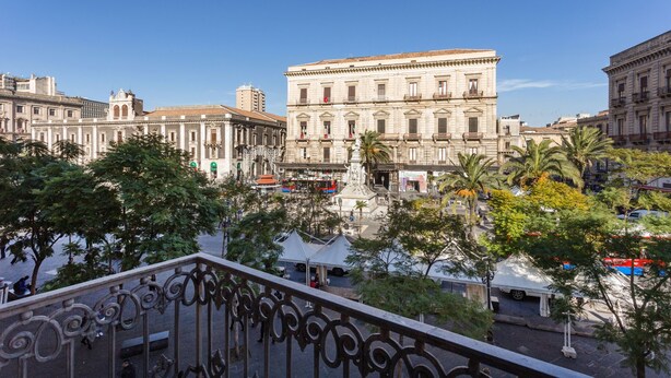 Un edificio histórico con arquitectura clásica, una plaza con palmeras y un cielo azul despejado.