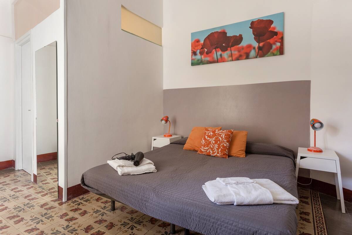 Un lit bien fait, recouvert d’une couverture grise, avec deux oreillers et une serviette blanche pliée posée dessus. Il y a deux tables de chevet avec des lampes, et un tableau représentant des fleurs rouges est accroché au mur au-dessus du lit.