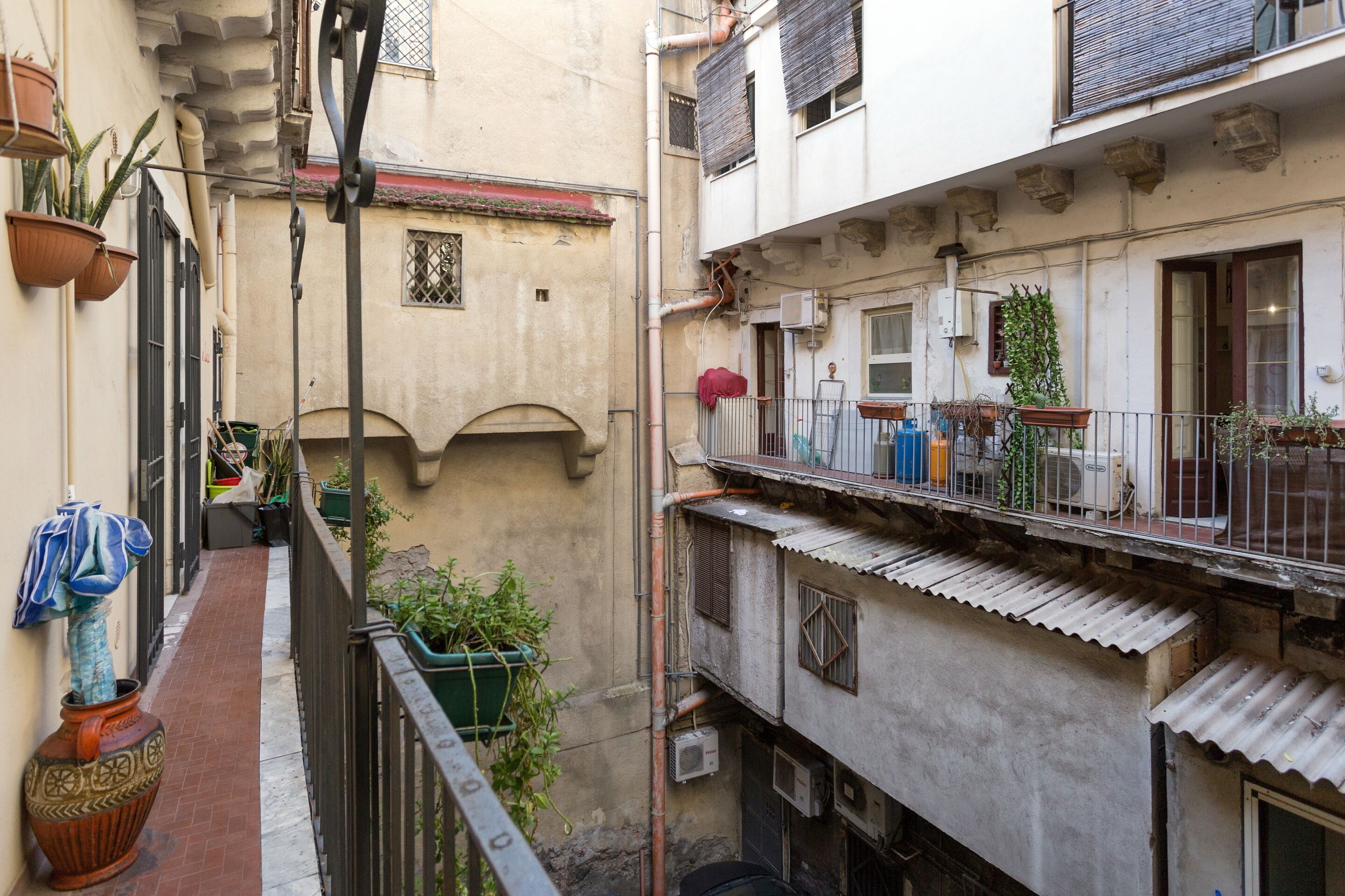 Une ruelle étroite, bordée de bâtiments des deux côtés, avec des balcons ornés de plantes en pot et un grand pot décoratif sur le côté gauche.