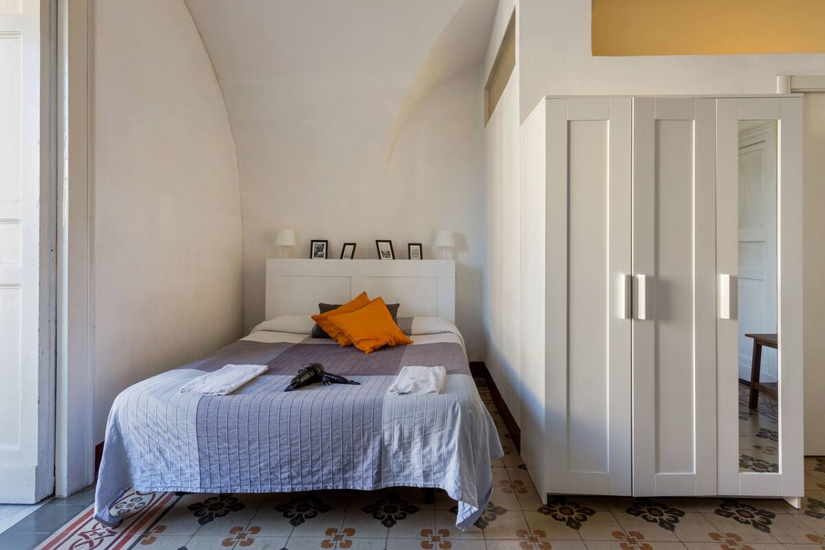 Un lit bien fait, recouvert d’une couvre-lit à rayures, avec un seul oreiller orange et un ensemble de tableaux encadrés accrochés au chevet.