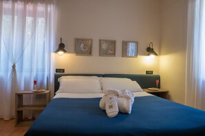 A neatly made bed with white linens, a blue bedspread, and a pair of white slippers placed on the bed. There are two bedside tables, each with a lamp and a glass of red wine. The room has a window with sheer curtains, and three framed pictures on the wall. - Villa Morgana Resort & Spa (Messina)