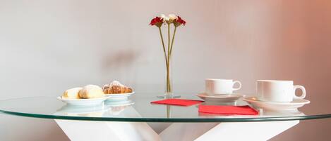 Una mesa con cubierta de vidrio y base blanca, un jarrón con flores, un plato de pasteles y dos tazas blancas sobre platillos.