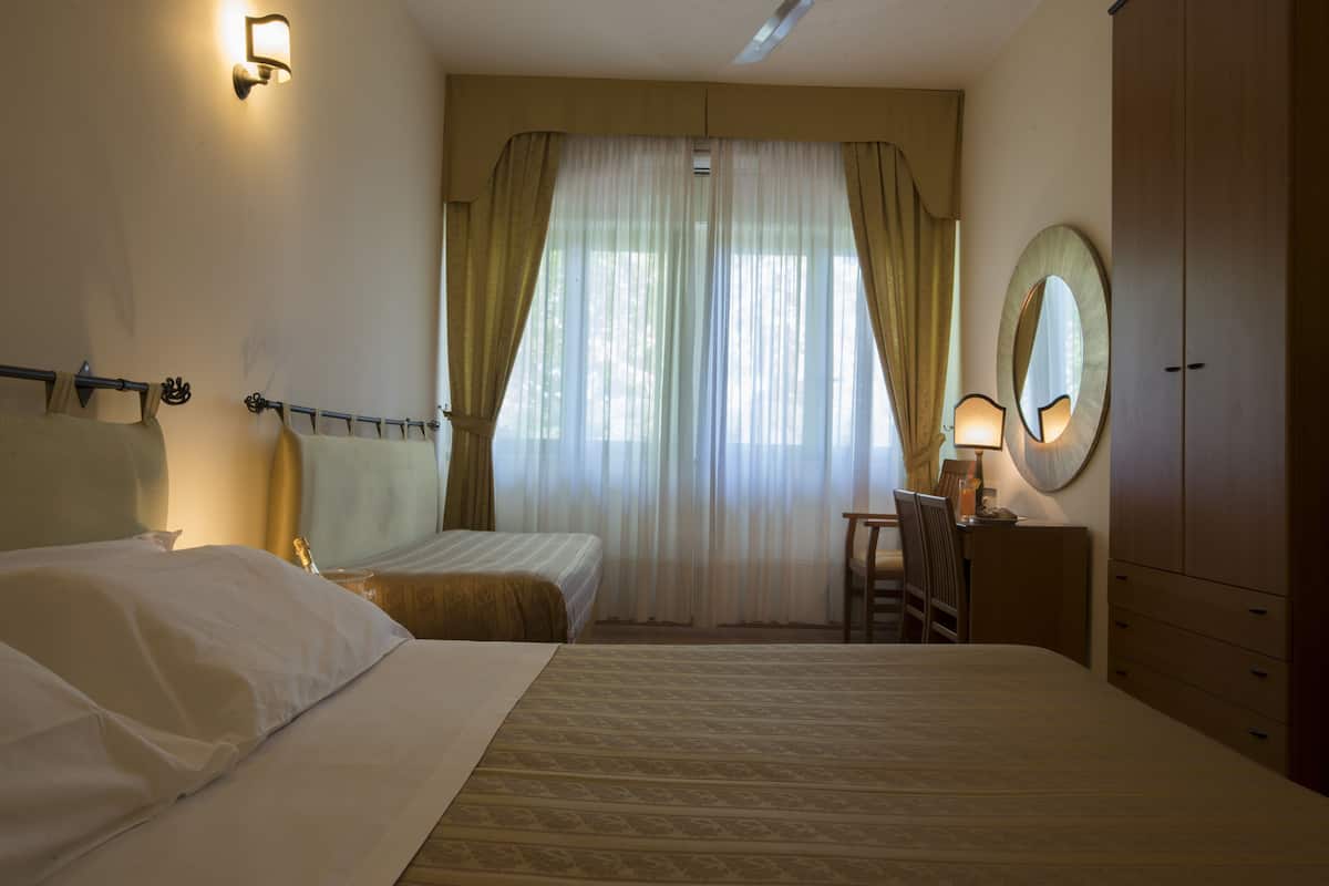 A hotel room with two beds, a desk with a chair, a wardrobe, and a large window with curtains.