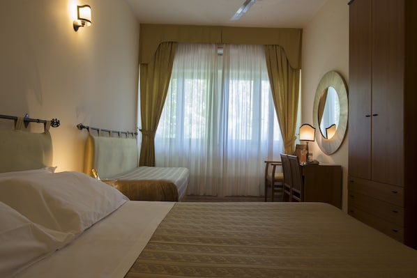 Habitación de hotel con dos camas, un escritorio con silla, un armario y un amplio ventanal con cortinas.