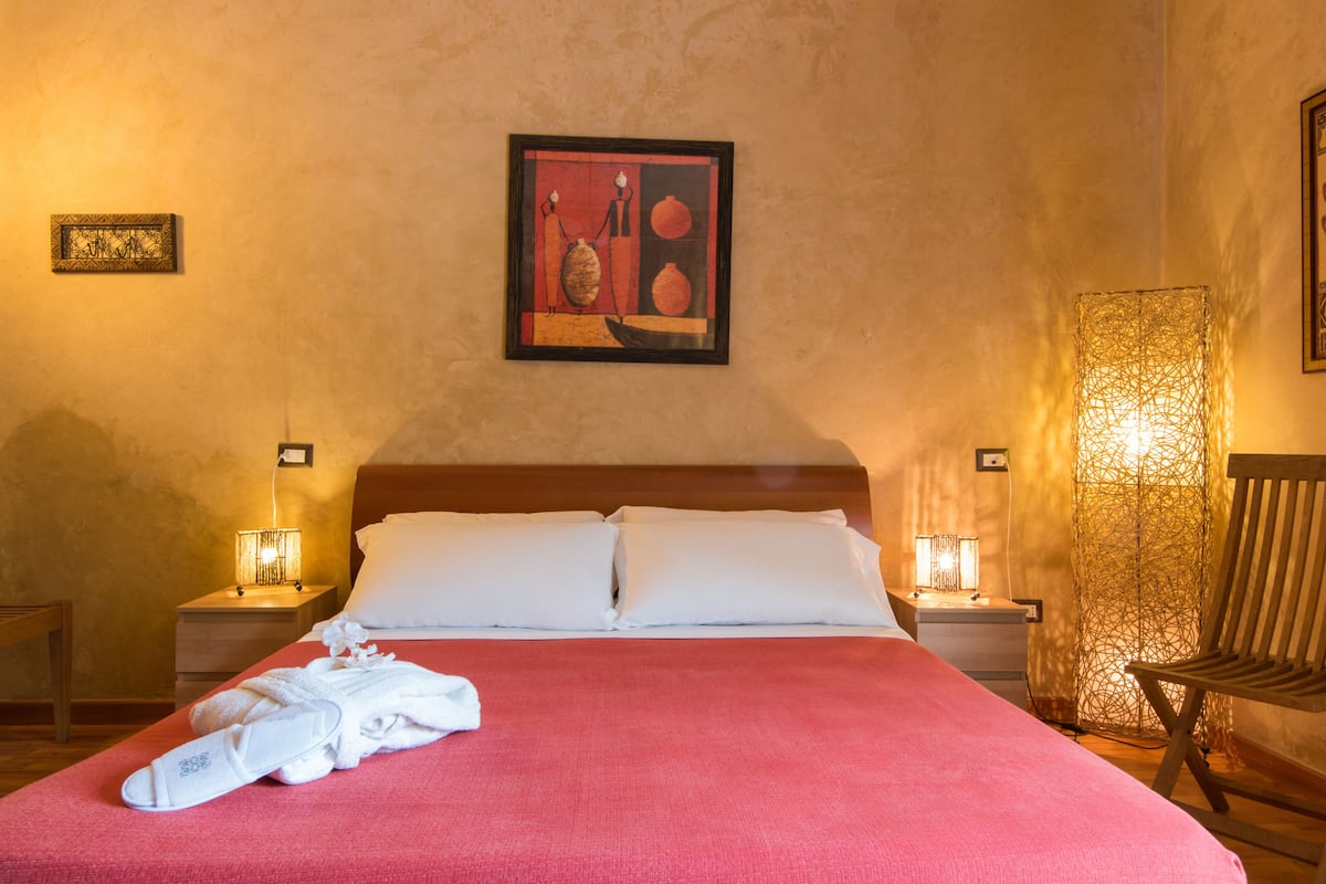 A neatly made bed with white linens, two bedside lamps, a framed artwork on the wall, and a wooden chair to the right.