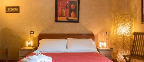 A neatly made bed with white linens, two bedside lamps, a framed artwork on the wall, and a wooden chair to the right.