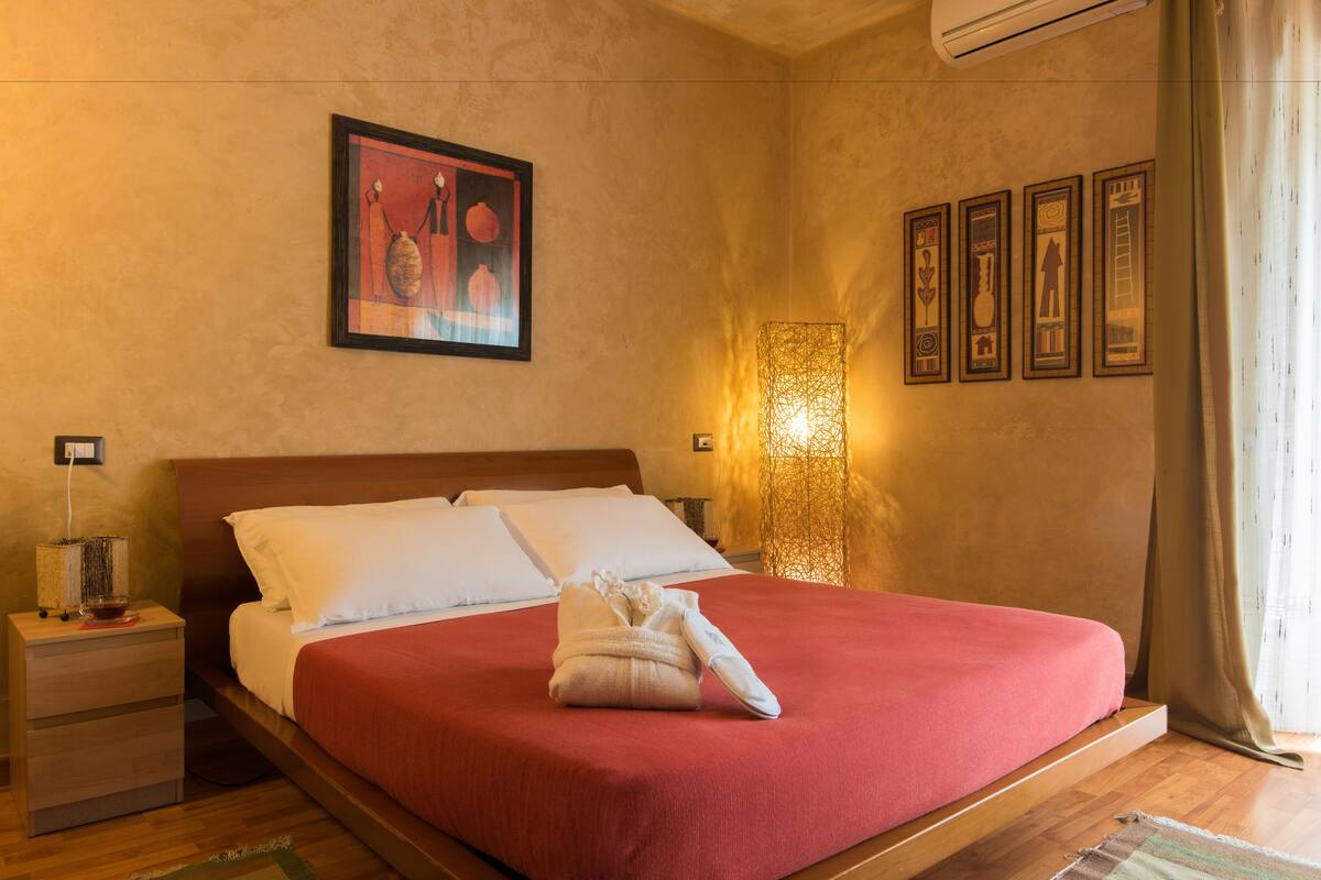 A neatly made bed with a red bedspread, a white pillow, and a towel on the side. There is a framed picture on the wall above the bed and a wall-mounted lamp to the right.