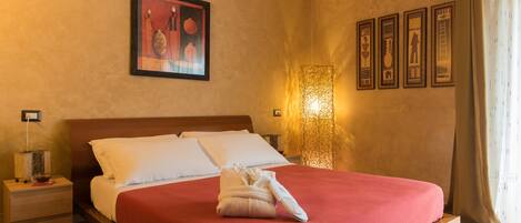 Una cama bien hecha con una colcha roja, una almohada blanca y una toalla al lado. Encima de la cama, hay un cuadro enmarcado en la pared y una lámpara de pared a la derecha.
