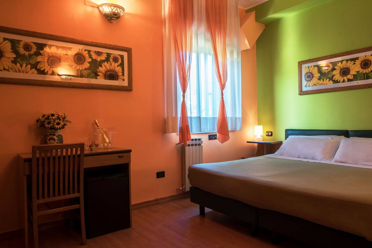 A bedroom with a bed, a desk with a chair, a radiator, and a window with sheer curtains.