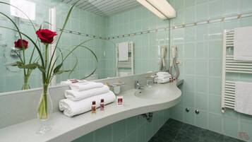 Une salle de bain avec un grand miroir, un lavabo et un porte-serviettes. Un vase contenant deux roses rouges est posé sur le comptoir.