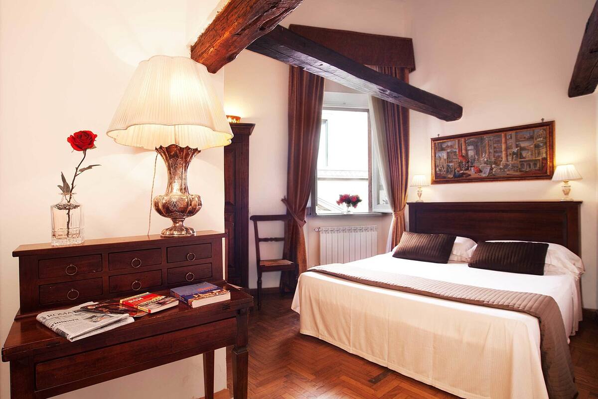A bedroom with a wooden bed, a bedside table with a lamp, a chair, and a window with curtains.