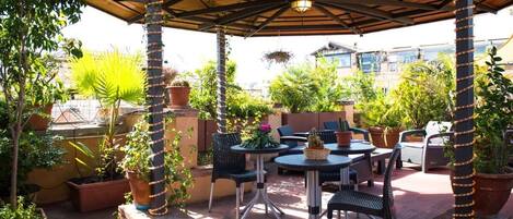 An outdoor patio with round tables and chairs, surrounded by potted plants and a pergola with string lights.