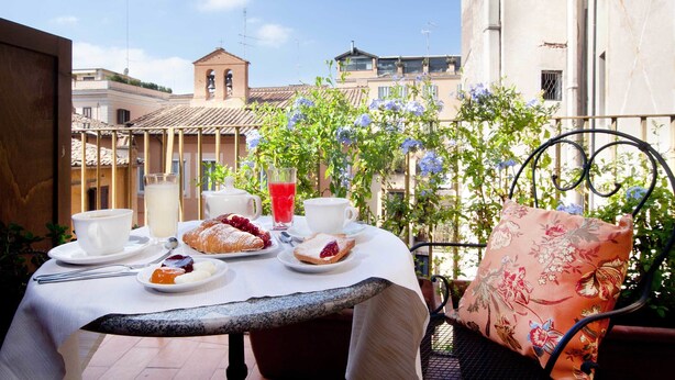 A table set with breakfast items, including pastries, a glass of juice, and a cup of coffee, on a balcony with a view of buildings and a bell tower. - Pantheon Inn (Rome)